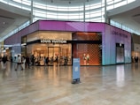 A modern, brightly lit Louis Vuitton storefront in a shopping mall with vibrant, colorful digital displays on the exterior. Shoppers can be seen walking in front of the store, and a few mannequins are positioned inside near the entrance. The store is surrounded by a spacious tiled floor with ample natural light coming through large windows and skylights.