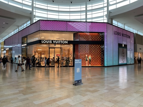 A modern, brightly lit Louis Vuitton storefront in a shopping mall with vibrant, colorful digital displays on the exterior. Shoppers can be seen walking in front of the store, and a few mannequins are positioned inside near the entrance. The store is surrounded by a spacious tiled floor with ample natural light coming through large windows and skylights.