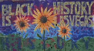 A vibrant mural featuring large sunflowers in the foreground with a deep blue sky and green hills in the background. The words 'BLACK IS HISTORY LAS VEGAS' are prominently displayed across the mural in bold, textured letters, creating a dynamic and thought-provoking composition.