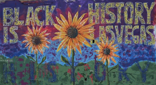 A vibrant mural featuring large sunflowers in the foreground with a deep blue sky and green hills in the background. The words 'BLACK IS HISTORY LAS VEGAS' are prominently displayed across the mural in bold, textured letters, creating a dynamic and thought-provoking composition.