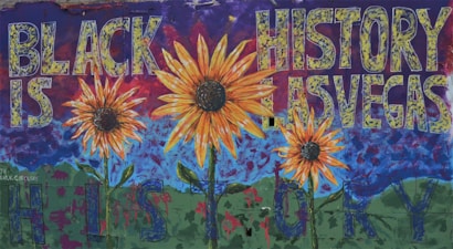 A vibrant mural featuring large sunflowers in the foreground with a deep blue sky and green hills in the background. The words 'BLACK IS HISTORY LAS VEGAS' are prominently displayed across the mural in bold, textured letters, creating a dynamic and thought-provoking composition.