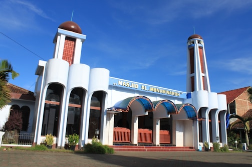 A mosque with modern architectural design featuring white multi-tiered columns, a clear blue sky in the background, and decorative blue arches above the entrance. The building includes tall minarets and some greenery at the base.