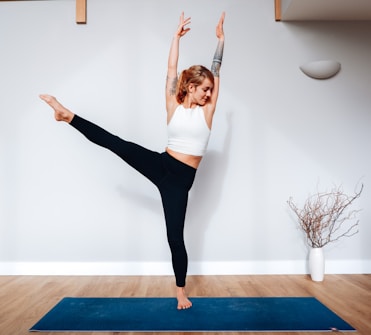 A person stands in a yoga pose on a blue mat, balancing on one foot with an extended leg and raised arms. The individual is wearing a white top and black leggings, and has visible tattoos. The setting is indoors with wooden flooring, a white wall, and a decorative vase with branches in the background.