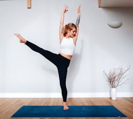 A person stands in a yoga pose on a blue mat, balancing on one foot with an extended leg and raised arms. The individual is wearing a white top and black leggings, and has visible tattoos. The setting is indoors with wooden flooring, a white wall, and a decorative vase with branches in the background.
