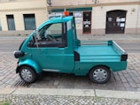 A small turquoise utility truck is parked on a cobblestone street. The truck has a single cab with two doors and a flatbed, suitable for light transport or deliveries. A small orange beacon light is mounted on the roof. The street is lined with old buildings, indicating an urban setting.