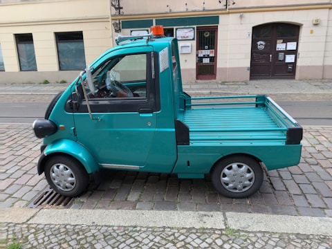 A small turquoise utility truck is parked on a cobblestone street. The truck has a single cab with two doors and a flatbed, suitable for light transport or deliveries. A small orange beacon light is mounted on the roof. The street is lined with old buildings, indicating an urban setting.