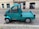A small turquoise utility truck is parked on a cobblestone street. The truck has a single cab with two doors and a flatbed, suitable for light transport or deliveries. A small orange beacon light is mounted on the roof. The street is lined with old buildings, indicating an urban setting.