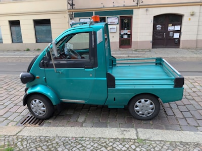 A small turquoise utility truck is parked on a cobblestone street. The truck has a single cab with two doors and a flatbed, suitable for light transport or deliveries. A small orange beacon light is mounted on the roof. The street is lined with old buildings, indicating an urban setting.