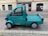 A small turquoise utility truck is parked on a cobblestone street. The truck has a single cab with two doors and a flatbed, suitable for light transport or deliveries. A small orange beacon light is mounted on the roof. The street is lined with old buildings, indicating an urban setting.