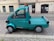 A small turquoise utility truck is parked on a cobblestone street. The truck has a single cab with two doors and a flatbed, suitable for light transport or deliveries. A small orange beacon light is mounted on the roof. The street is lined with old buildings, indicating an urban setting.