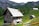 A rustic wooden cabin is situated in a mountainous area surrounded by lush greenery and other smaller wooden structures. The cabin features a slanted, shingle-covered roof, with a chimney and small windows. In the background, there are dense forests and towering rock formations.
