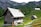 A rustic wooden cabin is situated in a mountainous area surrounded by lush greenery and other smaller wooden structures. The cabin features a slanted, shingle-covered roof, with a chimney and small windows. In the background, there are dense forests and towering rock formations.