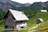 A rustic wooden cabin is situated in a mountainous area surrounded by lush greenery and other smaller wooden structures. The cabin features a slanted, shingle-covered roof, with a chimney and small windows. In the background, there are dense forests and towering rock formations.