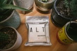 A package labeled 'ALT' is placed on a wooden table surrounded by various potted plants in ceramic and tin containers. The package indicates that it contains cannabis-infused liquid with THC content, and it has a minimalist design with a metallic finish.