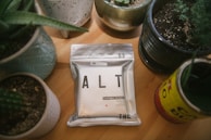 A package labeled 'ALT' is placed on a wooden table surrounded by various potted plants in ceramic and tin containers. The package indicates that it contains cannabis-infused liquid with THC content, and it has a minimalist design with a metallic finish.
