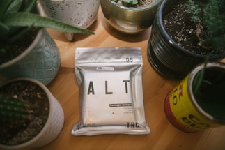 A package labeled 'ALT' is placed on a wooden table surrounded by various potted plants in ceramic and tin containers. The package indicates that it contains cannabis-infused liquid with THC content, and it has a minimalist design with a metallic finish.