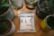 A package labeled 'ALT' is placed on a wooden table surrounded by various potted plants in ceramic and tin containers. The package indicates that it contains cannabis-infused liquid with THC content, and it has a minimalist design with a metallic finish.