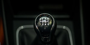 A close-up of a gear shift knob used for manual transmission in a car, featuring numbers and letters indicating the gear positions. The knob is shiny with a reflective surface, set against a dark background.