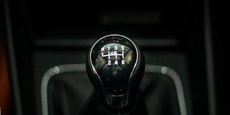 A close-up of a gear shift knob used for manual transmission in a car, featuring numbers and letters indicating the gear positions. The knob is shiny with a reflective surface, set against a dark background.