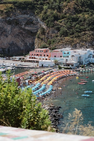 A coastal scene featuring a vibrant beach with rows of colorful umbrellas in shades of blue, orange, and yellow. The shoreline is populated with people enjoying the water and sunbathing. In the background, there is a small coastal town with pastel-colored buildings nestled against a rocky, vegetated hillside. The clear blue sky and calm ocean complete the picturesque setting.