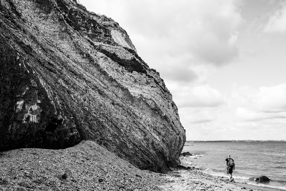 grayscale photo of person walking on beach near rock formation