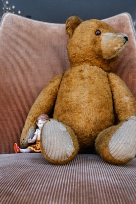 A large vintage teddy bear with a worn, classic appearance sits on a plush, corduroy chair. Nestled against the bear is a small porcelain doll, dressed in a floral skirt and knitted top, with red shoes and braided hair. The doll leans gently against the bear, creating a cozy and nostalgic scene.