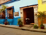 A vibrant street scene showcasing a colorful colonial building with bright blue and yellow walls. The entrance is adorned with decorative wooden window grilles and potted plants. A wooden door is open, revealing a rustic interior with a bull statue near the entrance.
