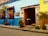 A vibrant street scene showcasing a colorful colonial building with bright blue and yellow walls. The entrance is adorned with decorative wooden window grilles and potted plants. A wooden door is open, revealing a rustic interior with a bull statue near the entrance.