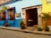 A vibrant street scene showcasing a colorful colonial building with bright blue and yellow walls. The entrance is adorned with decorative wooden window grilles and potted plants. A wooden door is open, revealing a rustic interior with a bull statue near the entrance.