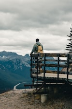 man in black jacket standing on brown wooden bench looking at mountains during daytime