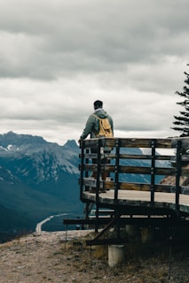 man in black jacket standing on brown wooden bench looking at mountains during daytime