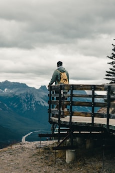 man in black jacket standing on brown wooden bench looking at mountains during daytime