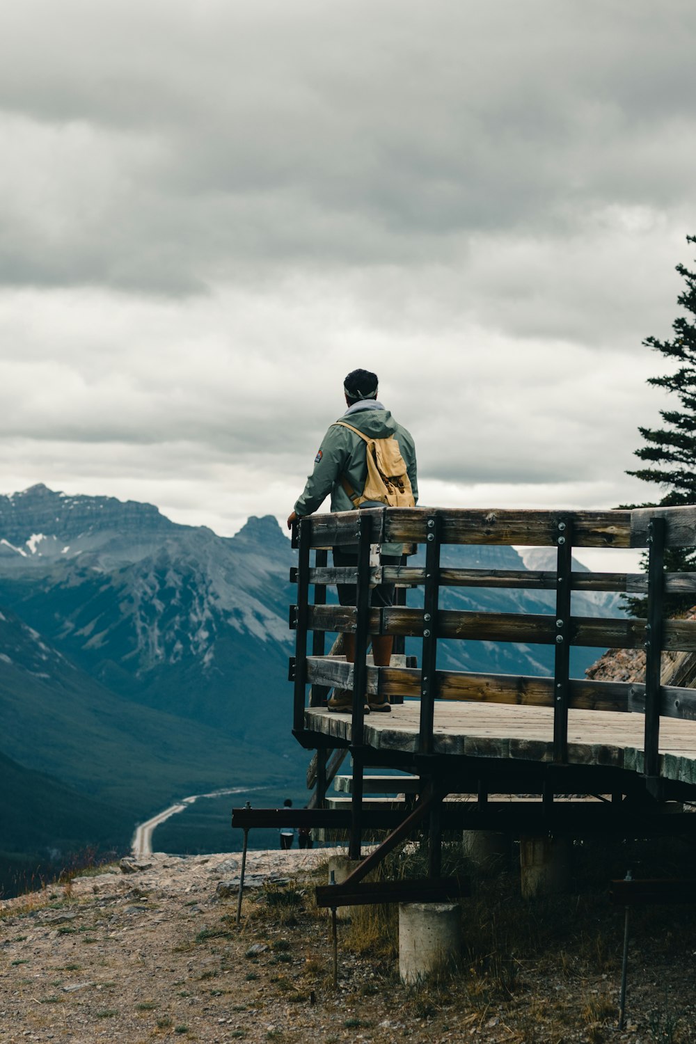 Hombre en chaqueta negra de pie en banco de madera marrón mirando las montañas durante el día