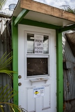 A doorway partially framed by green trim, featuring a sign that says 'PICK UP FOOD HERE' and another sign below with a QR code for scanning the menu. This is likely a service entrance for food pickup.