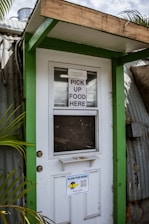 A doorway partially framed by green trim, featuring a sign that says 'PICK UP FOOD HERE' and another sign below with a QR code for scanning the menu. This is likely a service entrance for food pickup.