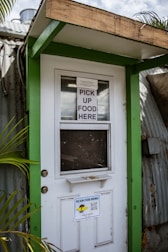 A doorway partially framed by green trim, featuring a sign that says 'PICK UP FOOD HERE' and another sign below with a QR code for scanning the menu. This is likely a service entrance for food pickup.