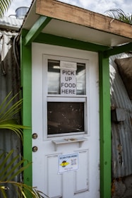 A doorway partially framed by green trim, featuring a sign that says 'PICK UP FOOD HERE' and another sign below with a QR code for scanning the menu. This is likely a service entrance for food pickup.