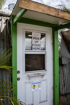 A doorway partially framed by green trim, featuring a sign that says 'PICK UP FOOD HERE' and another sign below with a QR code for scanning the menu. This is likely a service entrance for food pickup.
