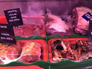 Various cuts of meat are displayed in a refrigerated section, including yearling minute steak and lamb roasts. The meat is placed on trays, with some pieces wrapped in plastic. Price tags are visible, indicating the cost per kilogram.