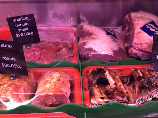 Various cuts of meat are displayed in a refrigerated section, including yearling minute steak and lamb roasts. The meat is placed on trays, with some pieces wrapped in plastic. Price tags are visible, indicating the cost per kilogram.
