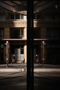 A business professional walks along a city street, his reflection visible in a large glass window. The building façade is modern with clean lines and large windows, and the glass reflects both the man and the urban environment around him.