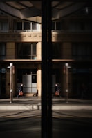 A business professional walks along a city street, his reflection visible in a large glass window. The building façade is modern with clean lines and large windows, and the glass reflects both the man and the urban environment around him.