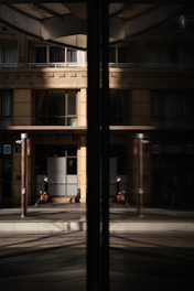 A business professional walks along a city street, his reflection visible in a large glass window. The building façade is modern with clean lines and large windows, and the glass reflects both the man and the urban environment around him.