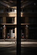 A business professional walks along a city street, his reflection visible in a large glass window. The building fa&ccedil;ade is modern with clean lines and large windows, and the glass reflects both the man and the urban environment around him.
