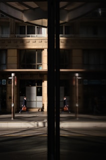 A business professional walks along a city street, his reflection visible in a large glass window. The building fa&ccedil;ade is modern with clean lines and large windows, and the glass reflects both the man and the urban environment around him.
