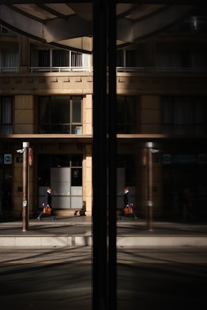 A business professional walks along a city street, his reflection visible in a large glass window. The building fa&ccedil;ade is modern with clean lines and large windows, and the glass reflects both the man and the urban environment around him.