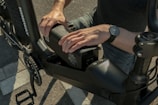 A person is installing or removing a battery pack from an electric bicycle. The individual is wearing a watch on the left wrist, and the bicycle features a robust frame. The setting appears to be outdoors on a paved surface.