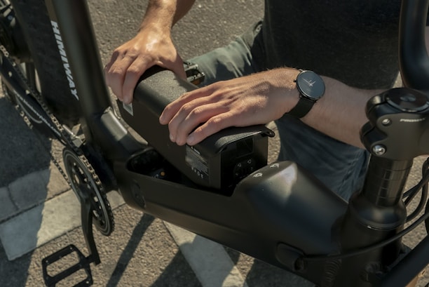 A person is installing or removing a battery pack from an electric bicycle. The individual is wearing a watch on the left wrist, and the bicycle features a robust frame. The setting appears to be outdoors on a paved surface.