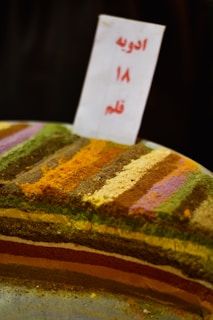 A display of various colorful spices arranged in horizontal layers. Each layer consists of a different spice, creating a vibrant and textured pattern. A sign with text in another language is placed upright in the center.