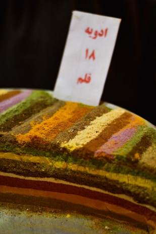 A display of various colorful spices arranged in horizontal layers. Each layer consists of a different spice, creating a vibrant and textured pattern. A sign with text in another language is placed upright in the center.